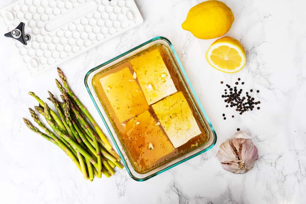 Four slabs of tofu in marinade in glass dish with black peppercorns, raw asparagus, garlic, EZ Tofu Press, and lemons off to side on white marble counter