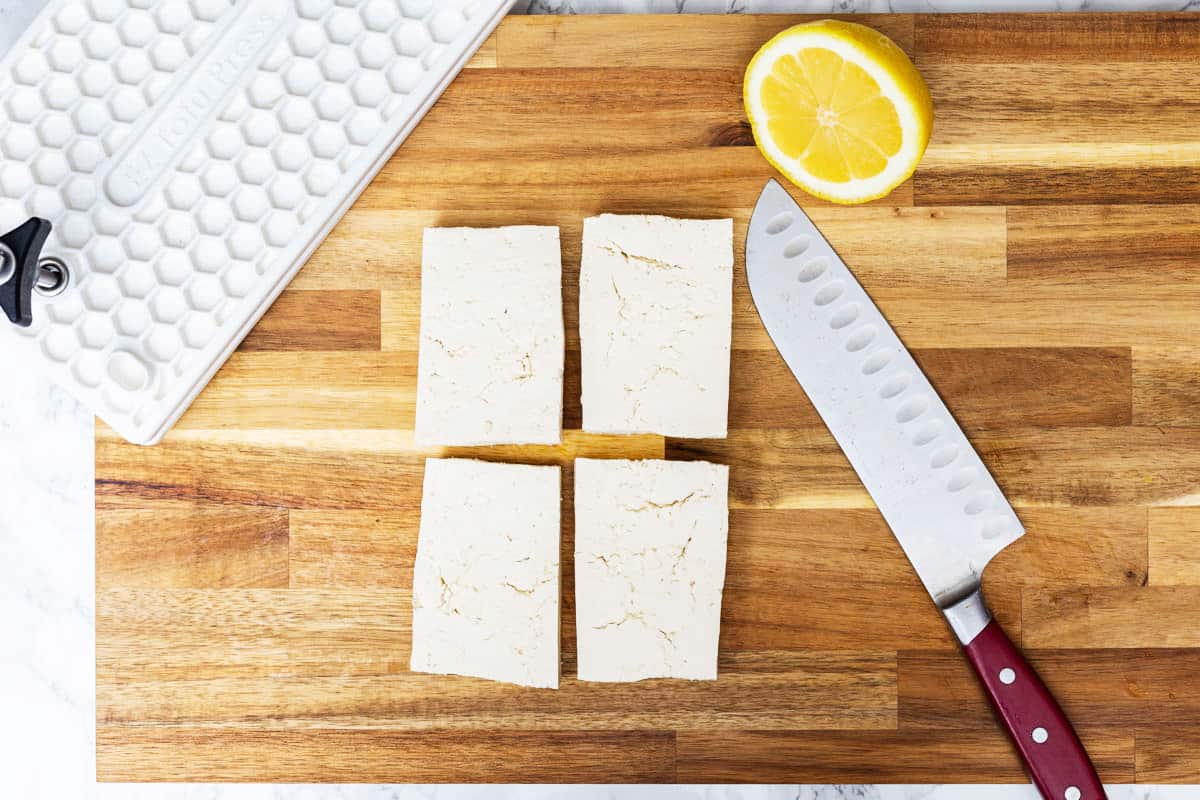 Four slabs of raw tofu on wooden cutting board with chef's knife, lemon half, and EZ Tofu Press off to side