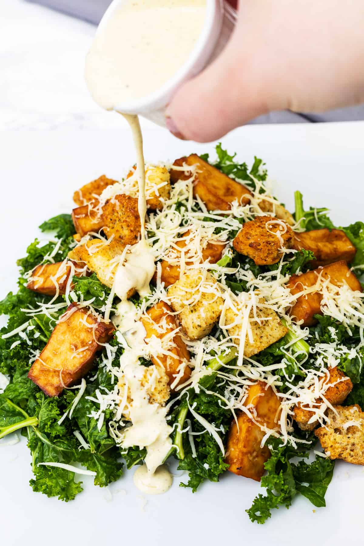 Hand pouring dressing onto plate of kale salad with croutons, tofu, and vegan cheese