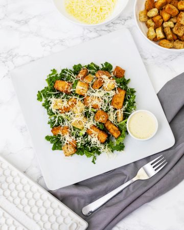 Plate of kale caesar salad with gray cloth napkin and fork surrounded by bowls of ingredients