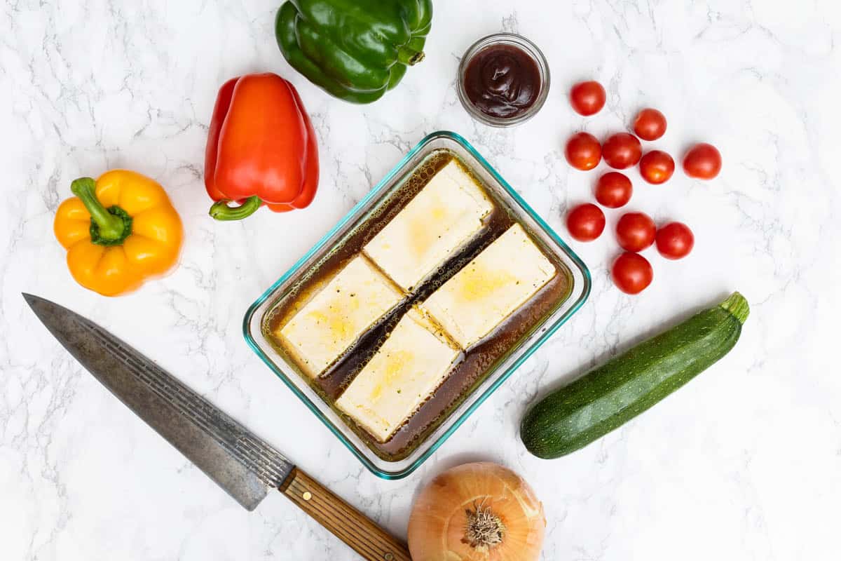 Four slabs of tofu marinating in shallow glass dish surrounded by bell peppers, tomatoes, barbecue sauce, zucchini, and onion on white marble counter
