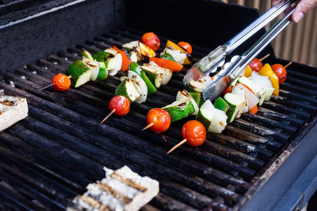 Hand turning grilled vegetable skewers with metal tongs on gas grill
