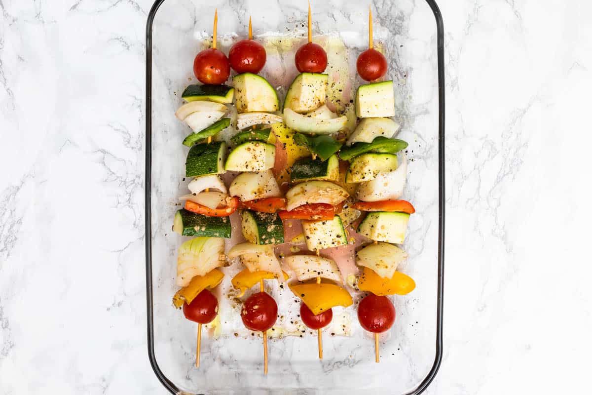 Vegetables on bamboo skewers marinating in glass dish on white marble surface