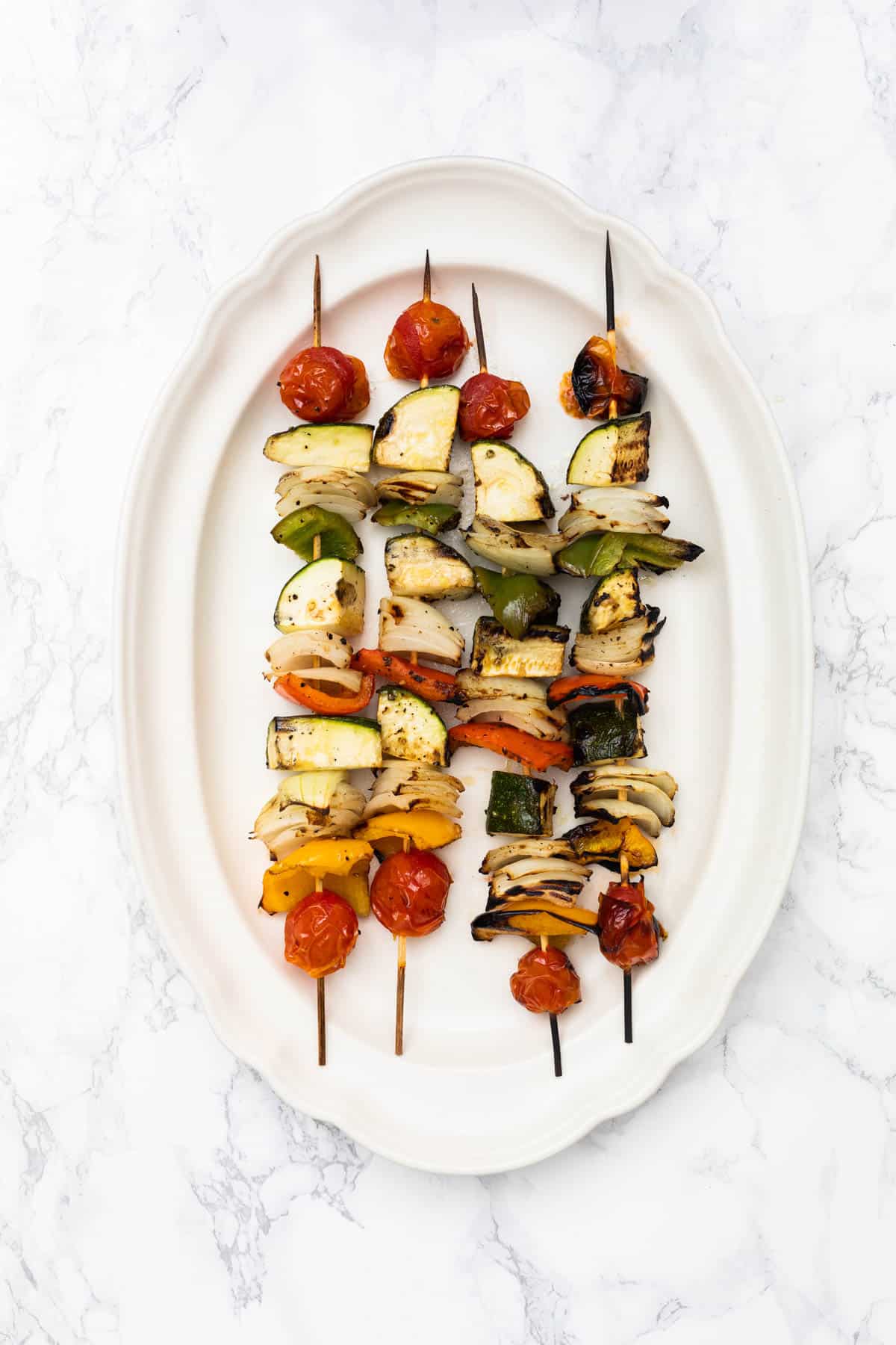 Four grilled vegetable skewers on plate on white marble counter