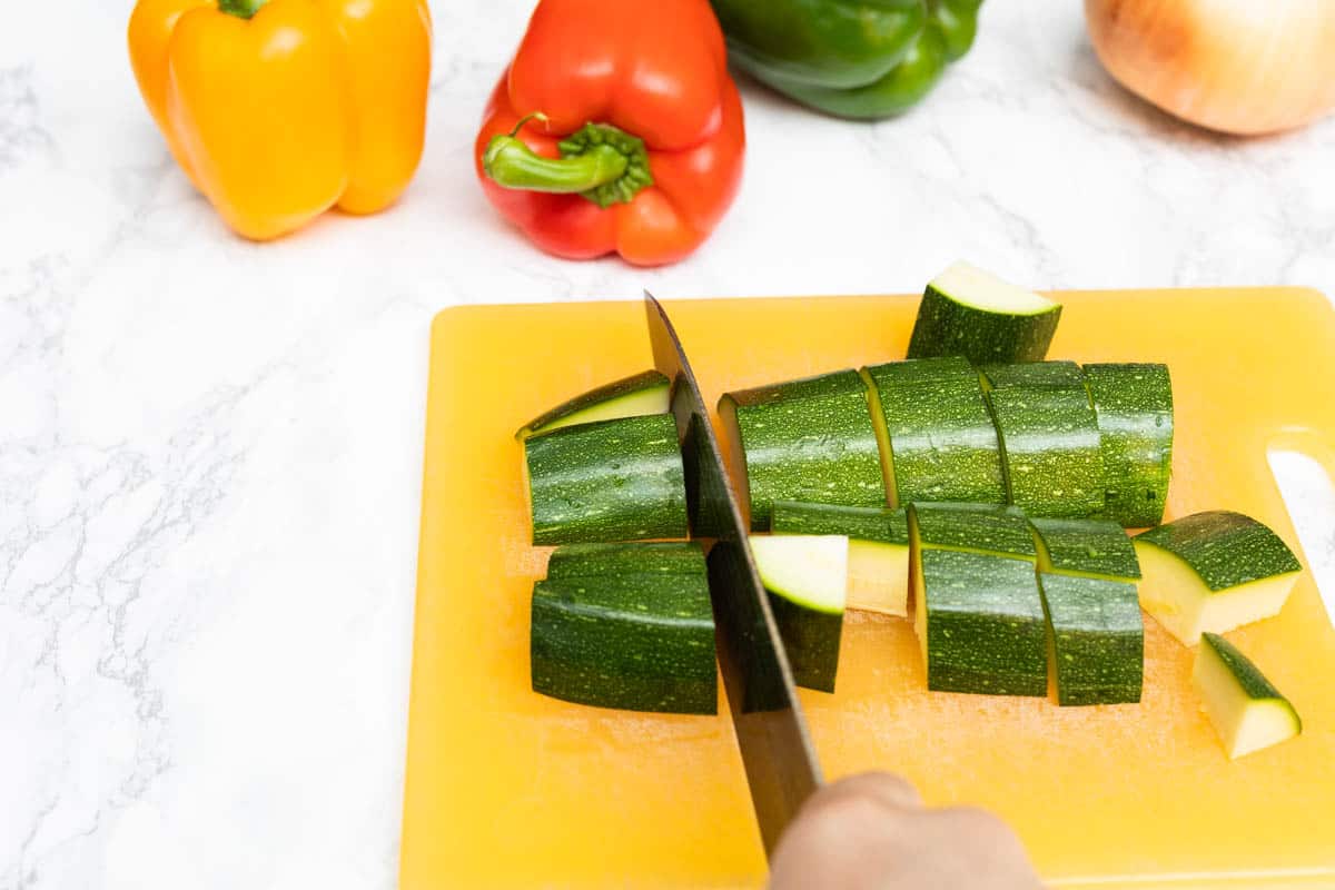 Hand chopping zucchini into large chunks with bell peppers and onion off to the side on white marble counter