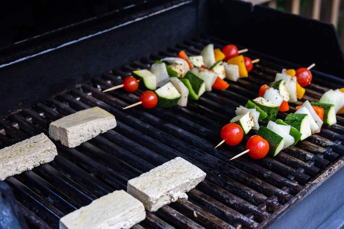 Four pieces of raw marinated tofu and four vegetable skewers on the grates in a gas grill