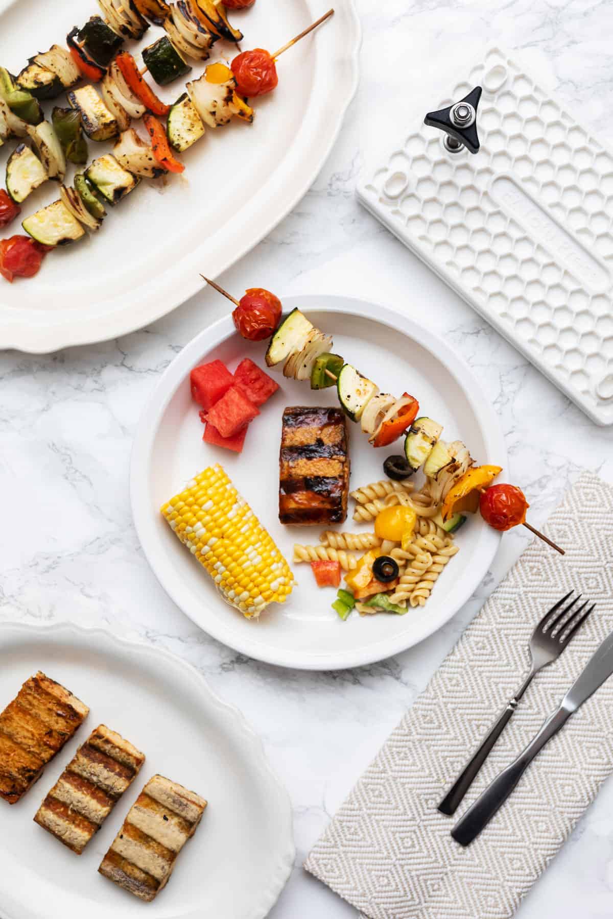 Plate with BBQ grilled tofu, vegetable skewer, pasta salad, corn on the cob, and watermelon with plates of food and place settings