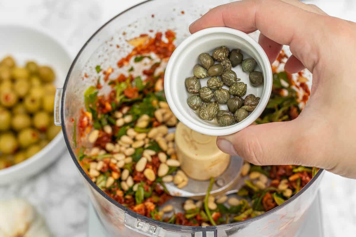 Hand pouring small bowl of capers into food processor container with sun-dried tomatoes, pine nuts, and fresh herbs