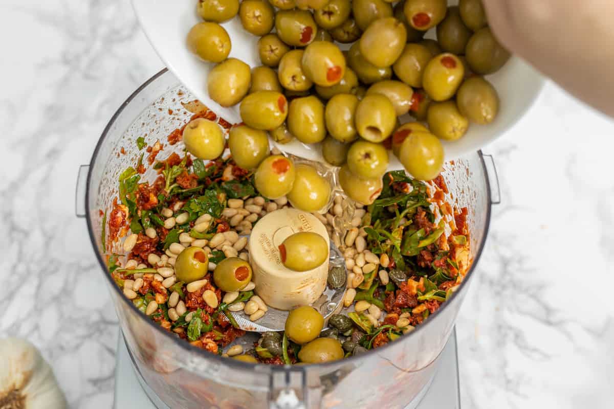 Hand pouring bowl of green olives into food processor container with sun-dried tomatoes, pine nuts, and fresh herbs